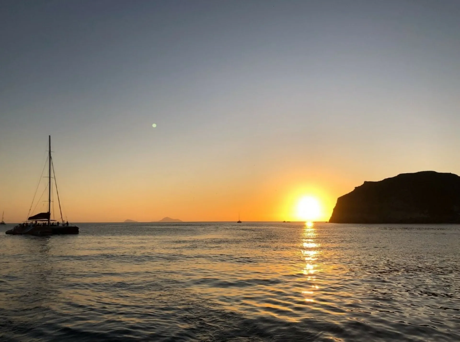 Sun setting beside a cliff off Santorini with a sailing boat on the water